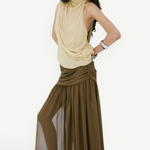 A person poses against a white wall wearing a pale yellow sleeveless top and an olive green Agapito Skirt that creates an elongated, floor-length chiffon silhouette.