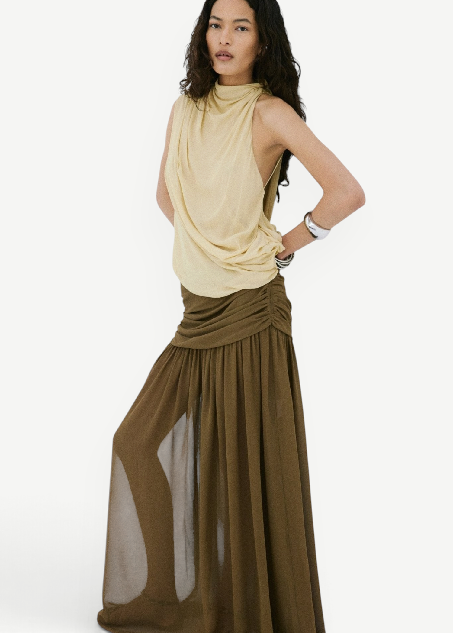 A person poses against a white wall wearing a pale yellow sleeveless top and an olive green Agapito Skirt that creates an elongated, floor-length chiffon silhouette.