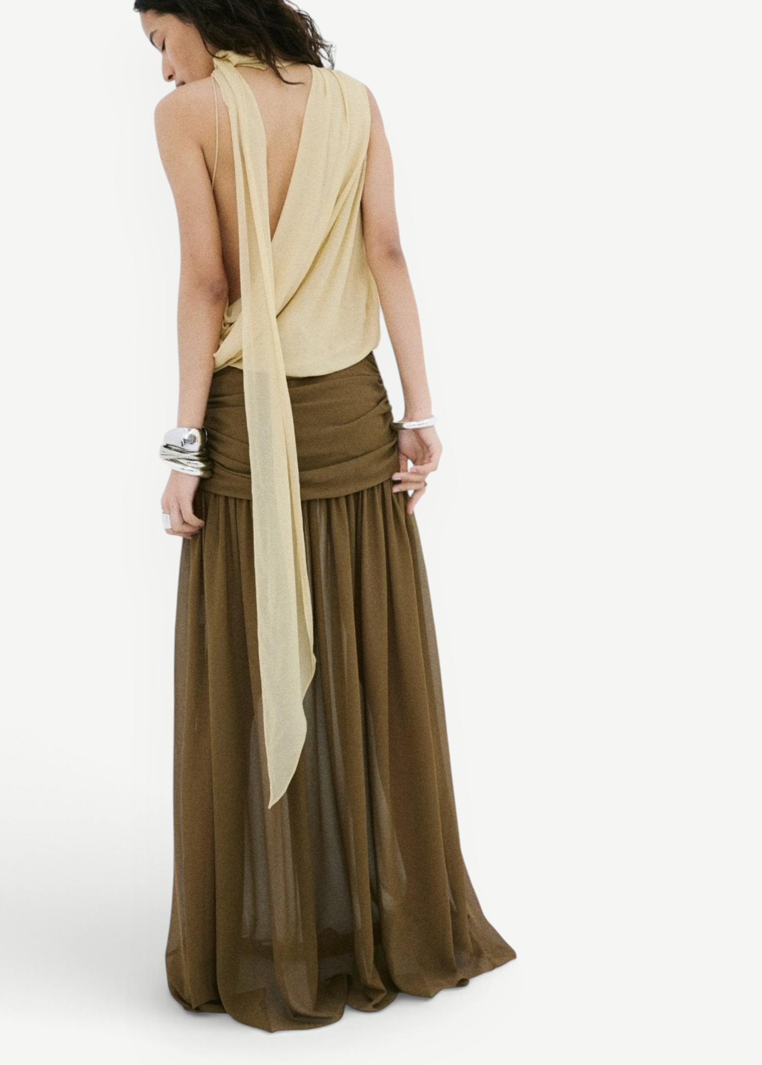 A person wearing a pale yellow, sleeveless, open-back top tucked into an Agapito Skirt, a long, taupe chiffon skirt with a gathered waistband that creates an elongated silhouette.