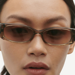 Close-up of a person wearing the Billy rectangular brown sunglasses, crafted from Mazzucchelli cellulose acetate, featuring dark straight hair pulled back, neutral makeup, and a serious expression, set against a plain light background.