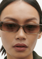 Close-up of a person wearing the Billy rectangular brown sunglasses, crafted from Mazzucchelli cellulose acetate, featuring dark straight hair pulled back, neutral makeup, and a serious expression, set against a plain light background.
