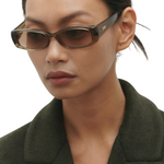 Wearing the Billy rectangular brown sunglasses, crafted from premium Mazzucchelli cellulose acetate, a woman with straight dark hair styled in a low bun and dressed in a dark green blazer looks down with a neutral expression against a plain light background.