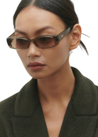 Wearing the Billy rectangular brown sunglasses, crafted from premium Mazzucchelli cellulose acetate, a woman with straight dark hair styled in a low bun and dressed in a dark green blazer looks down with a neutral expression against a plain light background.