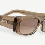 Close-up of the Billy sunglasses, showcasing a thick, rectangular frame in brown crafted from premium Mazzucchelli cellulose acetate. The metallic DMY logo adorns the temple, while the tinted lenses offer UV protection. Set against a plain white background.