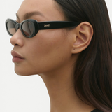 Model wearing KABE SHOP INC Naomi oval black Italian acetate sunglasses in a profile view against a white background.
