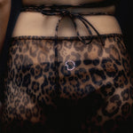 A close-up of a person wearing the Naj Skirt—a sheer, leopard-pattern summer maxi-skirt tied at the waist, featuring a small rhinestone detail at the center back. The background is dark.