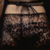 A close-up of a person wearing the Naj Skirt—a sheer, leopard-pattern summer maxi-skirt tied at the waist, featuring a small rhinestone detail at the center back. The background is dark.