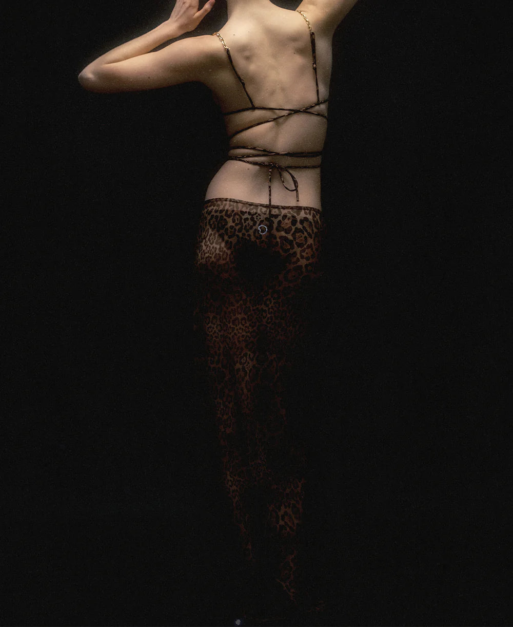 A person stands with their back to the camera in dim lighting, wearing a strappy top and the Naj Skirt—a summer maxi-skirt featuring a bold leopard pattern—with one arm raised and the other bent behind their head.