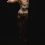 A person stands with their back to the camera in dim lighting, wearing a strappy top and the Naj Skirt—a summer maxi-skirt featuring a bold leopard pattern—with one arm raised and the other bent behind their head.