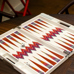 An open ZigZag Backgammon Board Game with a colorful design sits on a wooden coffee table. The set’s vibrant acrylic accents catch the light, while two dice rest on the board and part of a red chair is visible in the background.