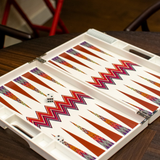 An open ZigZag Backgammon Board Game with a colorful design sits on a wooden coffee table. The set’s vibrant acrylic accents catch the light, while two dice rest on the board and part of a red chair is visible in the background.