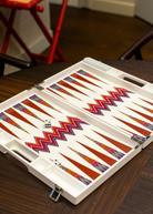 An open ZigZag Backgammon Board Game with a colorful design sits on a wooden coffee table. The set’s vibrant acrylic accents catch the light, while two dice rest on the board and part of a red chair is visible in the background.