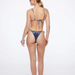 A woman stands with her back to the camera against a plain white background, wearing the blue patterned Praia Bottom bikini with adjustable straps and sunglasses. Her hair is styled in a low ponytail, she holds a white accessory, and wears sandals.