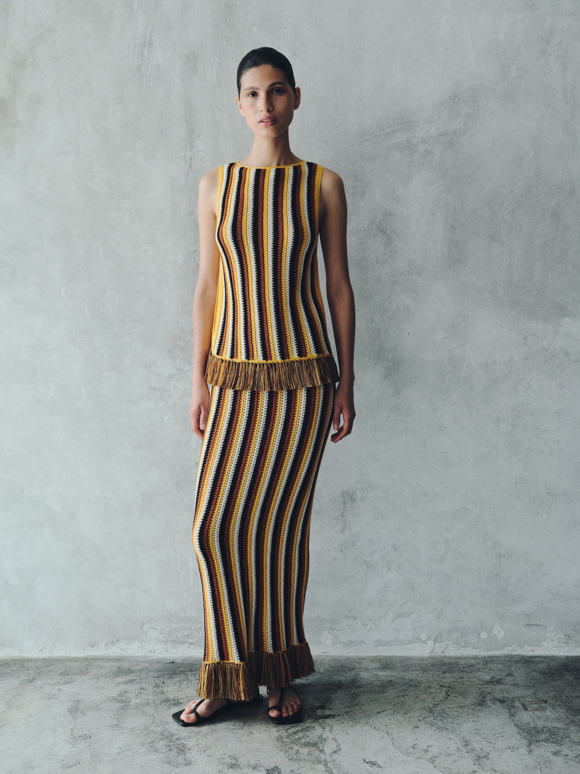 A woman wears a Misti Sleeveless Top paired with a matching maxi skirt featuring vertical stripes and fringed hems.