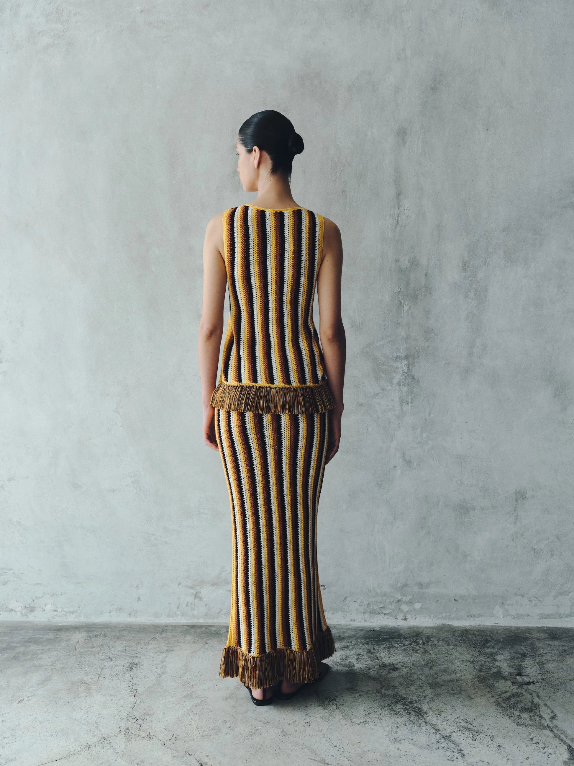Back view of a person wearing a Misti Sleeveless Top and matching long skirt, both featuring vertical stripes in brown, yellow, and white with artisanal detailing at the fringe hems.