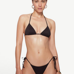 A woman with light skin and straight brown hair pulled back stands against a plain background, wearing the eco-friendly Praia Bottom black bikini with triangle bikini bottoms and gold hoop earrings. She has a neutral expression.