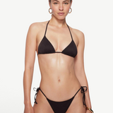 A woman with light skin and straight brown hair pulled back stands against a plain background, wearing the eco-friendly Praia Bottom black bikini with triangle bikini bottoms and gold hoop earrings. She has a neutral expression.