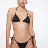 A woman with light skin and straight brown hair pulled back stands against a plain background, wearing the eco-friendly Praia Bottom black bikini with triangle bikini bottoms and gold hoop earrings. She has a neutral expression.