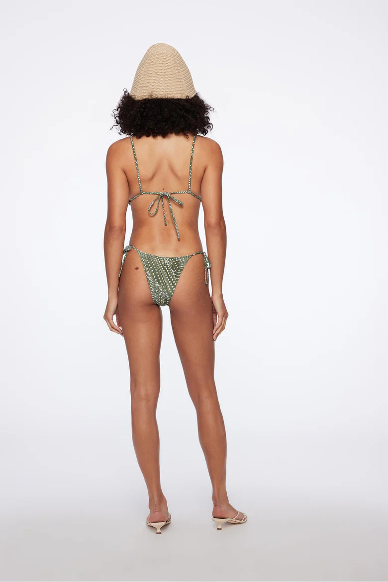 A woman stands barefoot, facing away from the camera against a plain white background, wearing the Equator Top Swimwear—a green patterned triangle bikini top made from recycled nylon—and a woven hat.