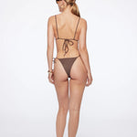 A woman stands facing away, wearing the Praia Bottom—a brown triangle bikini bottom crafted from eco-friendly materials like ECONYL® regenerated nylon. She accessorizes with sunglasses, hoop earrings, a bracelet, and flip-flops. Her hair is tied back against a plain white background.