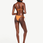 A woman with braided hair stands facing away, wearing the eco-friendly Praia Bottom—an orange triangle bikini bottom crafted from Peach Perfect fabric—paired with heeled sandals against a plain white background. This sustainable swimwear look is as stylish as it is mindful of the environment.