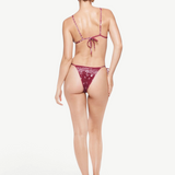 A woman stands facing away, wearing the Praia Bottom—a maroon bikini bottom with a bandana print and adjustable straps—paired with heeled sandals, against a plain white background.
