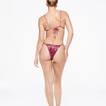 A woman stands facing away, wearing the Praia Bottom—a maroon bikini bottom with a bandana print and adjustable straps—paired with heeled sandals, against a plain white background.