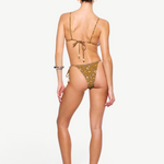 A woman stands facing away, wearing the Praia Bottom—a brown floral tie bikini bottom crafted from recycled nylon swimwear—paired with sandals. Her hair is tied back, and she has a bracelet on her left wrist. The background is plain white.