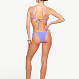 A woman stands with her back to the camera against a white background, wearing a lavender Praia Bottom—an adjustable tie bikini bottom—paired with sandals. Her hair is tied back, and she accessorizes with sunglasses and a bracelet, posing with one hand on her hip.