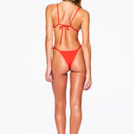 A woman stands facing away, wearing the Praia Bottom—a red bikini with high-cut, adjustable side-tie bottoms and thin straps. She has long, wavy hair and is barefoot on a plain white background.