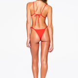 A woman stands facing away, wearing the Praia Bottom—a red bikini with high-cut, adjustable side-tie bottoms and thin straps. She has long, wavy hair and is barefoot on a plain white background.