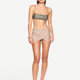 A woman stands against a white background wearing a snakeskin-patterned crop top, the Lumiere Skirt—a beige sheer wrap mini skirt—black sandals, and bracelets, with straight dark hair tucked behind her ears.