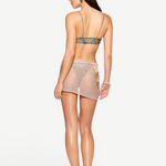 A woman stands facing away, showcasing the Lumiere Skirt—a sheer wrap mini skirt embellished with sequin bead accents. She pairs it with a strappy bikini top and high-heeled sandals, while her long brown hair cascades down her back. A chunky bracelet adorns her left wrist, all set against a plain white background.