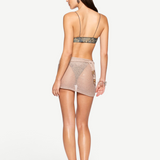 A woman stands facing away, showcasing the Lumiere Skirt—a sheer wrap mini skirt embellished with sequin bead accents. She pairs it with a strappy bikini top and high-heeled sandals, while her long brown hair cascades down her back. A chunky bracelet adorns her left wrist, all set against a plain white background.