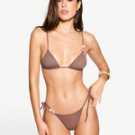 A woman with long brown hair wears the Praia Bottom, a brown bikini bottom crafted from eco-friendly swimwear materials and featuring adjustable tie sides with bead accents, standing against a plain white background.