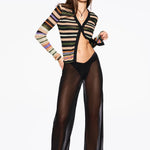 A woman stands confidently against a plain white background wearing black sheer pants over black underwear, the Linear Cardigan—a multi-colored striped rib knitted cardigan—paired with black sandals, sunglasses, and jewelry.