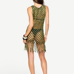A woman stands facing away against a plain white background, wearing the Castaway Dress—a sleeveless green and black macrame tunic with beaded fringe at the hem—paired with black sandals and black bangles.