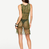 A woman stands facing away against a plain white background, wearing the Castaway Dress—a sleeveless green and black macrame tunic with beaded fringe at the hem—paired with black sandals and black bangles.