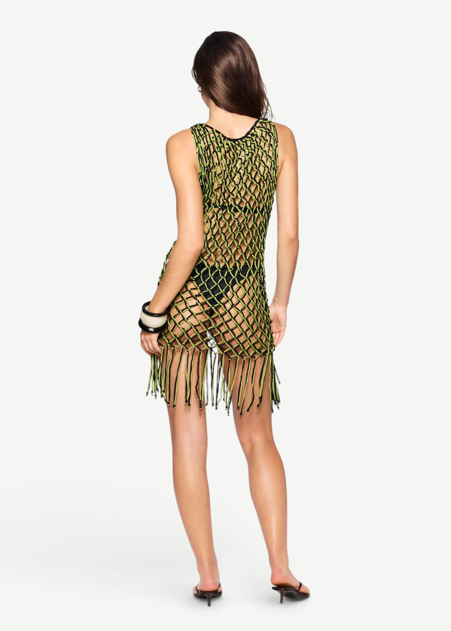 A woman stands facing away against a plain white background, wearing the Castaway Dress—a sleeveless green and black macrame tunic with beaded fringe at the hem—paired with black sandals and black bangles.