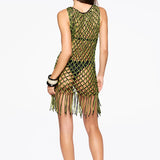 A woman stands facing away against a plain white background, wearing the Castaway Dress—a sleeveless green and black macrame tunic with beaded fringe at the hem—paired with black sandals and black bangles.