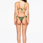 A woman stands facing away, wearing the Praia Bottom bikini in green with adjustable side ties and black heeled sandals against a plain white background. Her long, straight hair falls down her back and her arms rest relaxed at her sides.