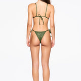 A woman stands facing away, wearing the Praia Bottom bikini in green with adjustable side ties and black heeled sandals against a plain white background. Her long, straight hair falls down her back and her arms rest relaxed at her sides.