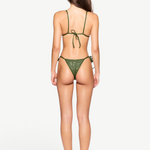 A woman stands facing away, wearing the Praia Bottom bikini in green with adjustable side ties and black heeled sandals against a plain white background. Her long, straight hair falls down her back and her arms rest relaxed at her sides.