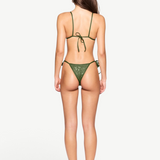 A woman stands facing away, wearing the Praia Bottom bikini in green with adjustable side ties and black heeled sandals against a plain white background. Her long, straight hair falls down her back and her arms rest relaxed at her sides.