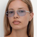 A person with straight blonde hair wears the Evangeline stainless steel frame, blue-tinted rectangular sunglasses and a white top, looking at the camera against a plain white background.