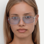 A young woman with straight, light brown hair wears the Evangeline sunglasses—light blue-tinted aviators with a sleek stainless steel frame—paired with small hoop earrings, looking directly at the camera with a neutral expression against a plain white background.