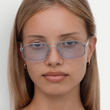 A young woman with straight, light brown hair wears the Evangeline sunglasses—light blue-tinted aviators with a sleek stainless steel frame—paired with small hoop earrings, looking directly at the camera with a neutral expression against a plain white background.