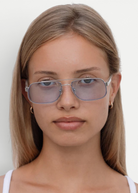 A young woman with straight, light brown hair wears the Evangeline sunglasses—light blue-tinted aviators with a sleek stainless steel frame—paired with small hoop earrings, looking directly at the camera with a neutral expression against a plain white background.