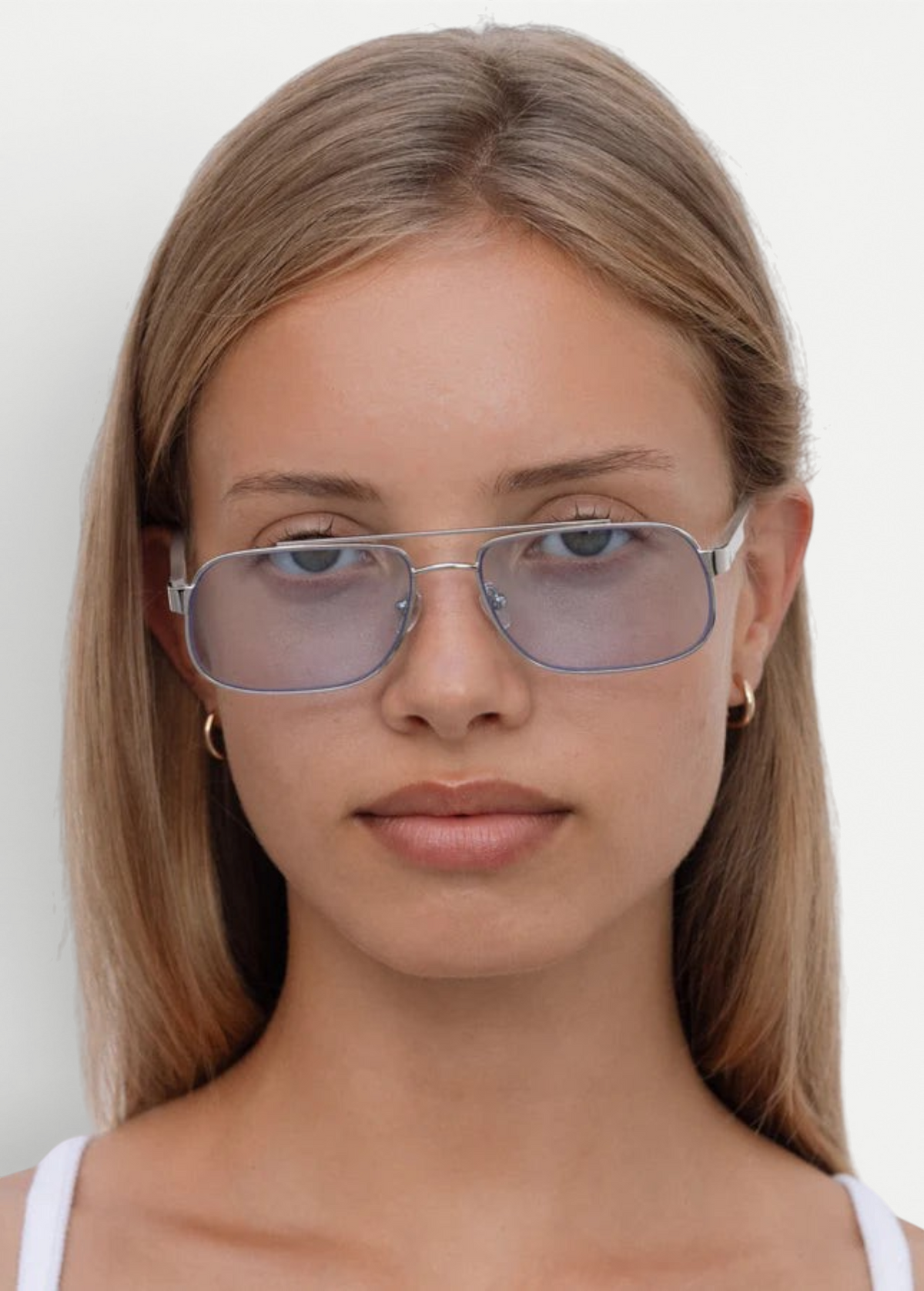 A young woman with straight, light brown hair wears the Evangeline sunglasses—light blue-tinted aviators with a sleek stainless steel frame—paired with small hoop earrings, looking directly at the camera with a neutral expression against a plain white background.