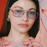 A woman with straight dark hair poses with one arm raised against a red background, wearing the Evangeline blue-tinted aviator sunglasses with a stainless steel frame, silver hoop earrings, and a pink patterned top.